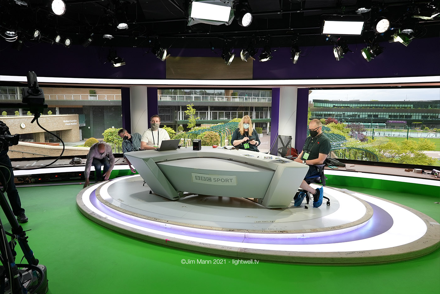 The set is shown whilst under construction. Plastic protective coverings are applied to almost all the surfaces. The BBC logo on the front of the desk is a paper mock-up for testing. The extent of the green floor can be seen and the real ceiling, a patchwork of lights and gridwork can be seen.