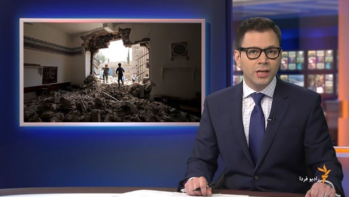 A male presenter in front of a virtual studio scene with a virtual creen displaying news footage behind his right shoulder