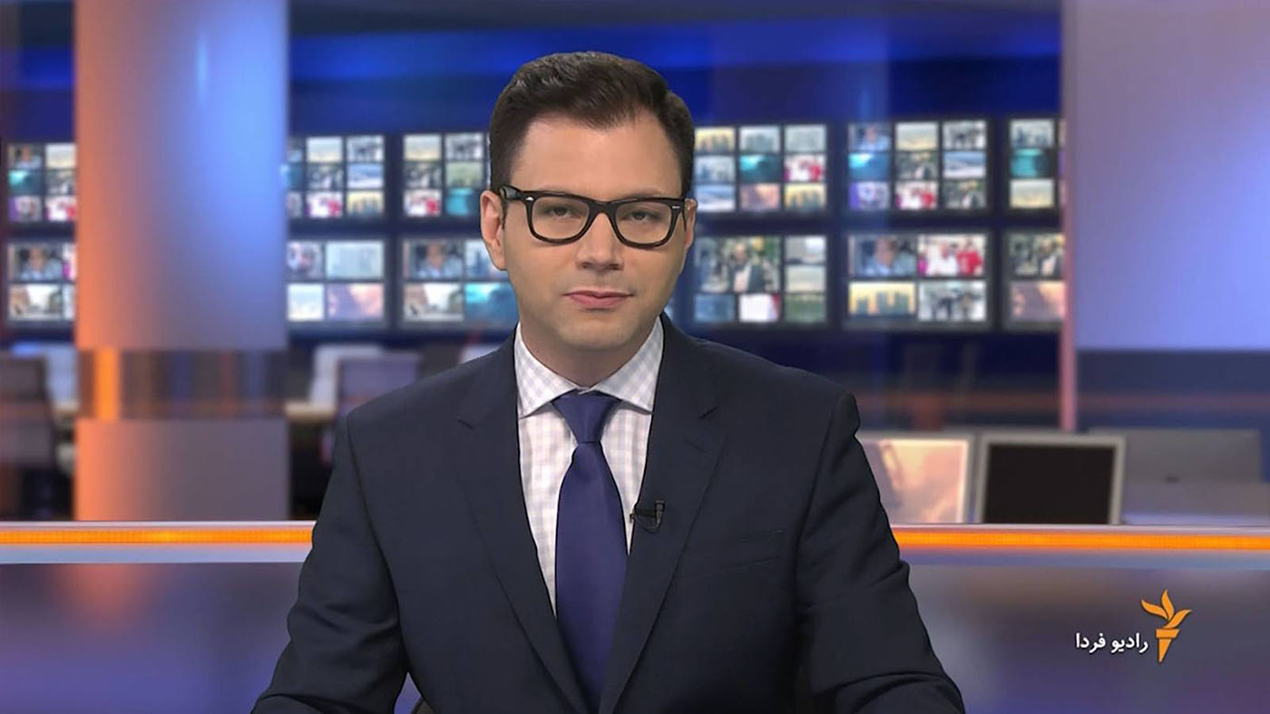 A male newsreader reads to the camera. In the background we see a virtual ewsrrom scene with architectural details and animated clips in the monitors.