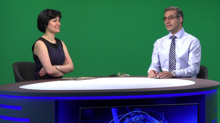 Two presenters sit at the desk. In the background, we see the greenscreen