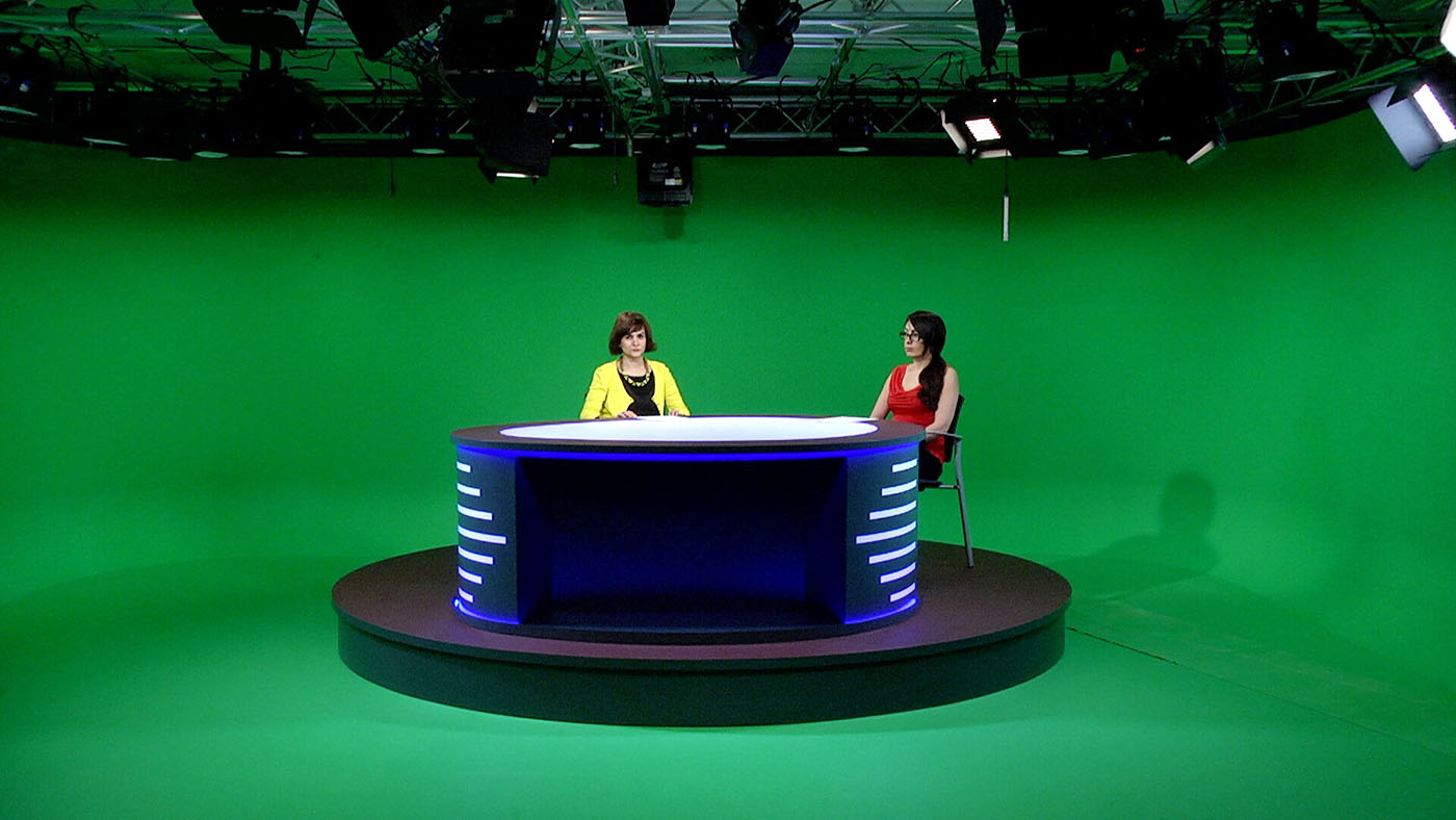 The desk with two stand-in presenters within the greenscreen studio. The lighting grid is visible above the desk, illustrating the limited height available within the real studio