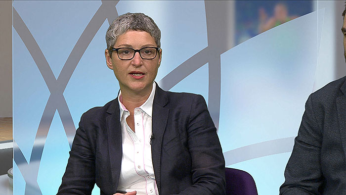 A short haired female presenter sits in fron of a frosted glass flower pattern.