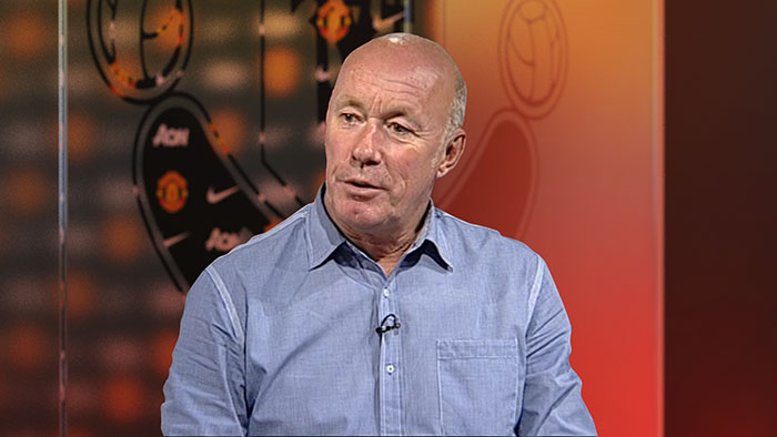 a mid-shot of Sammy McIlroy in front of a glass panel with a frosted MUFC logo behind.