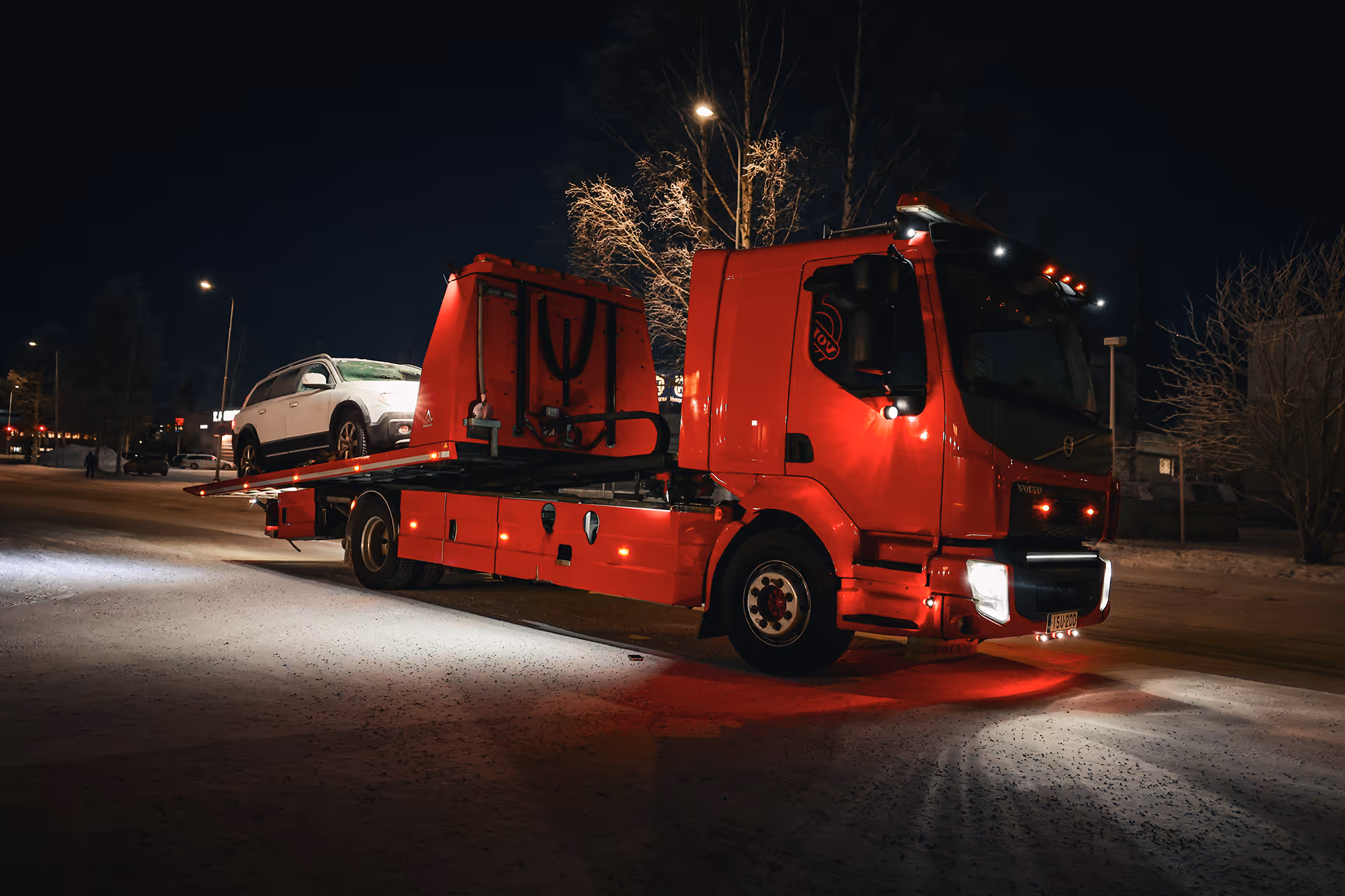 Punainen hinausauto yöaikaan, jossa valkoinen auto lavalla.