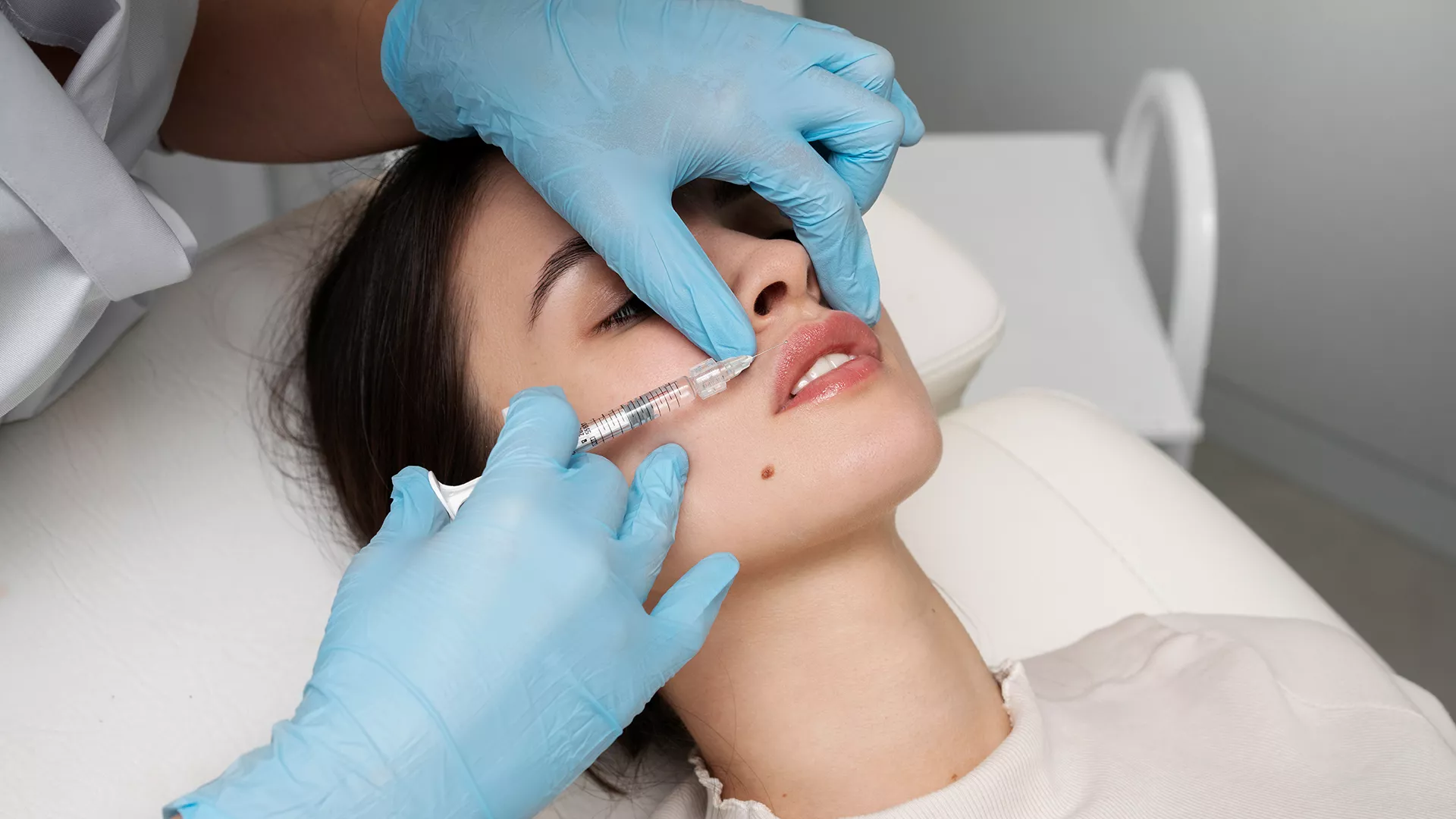 A patient receiving a cosmetic injectable treatment around the upper lip while a clinician wears protective gloves and uses a fine syringe in a clinical setting. For a Dental Examination, key related areas include oral health assessment, tooth decay detection, gum health evaluation, oral cancer screening and preventive dentistry, helping identify dental concerns early and guide personalised treatment planning for long-term oral health.