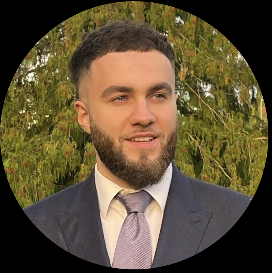 Smiling man with short curly hair and beard wearing a dark suit, light shirt, and lavender tie standing outdoors with green foliage background.