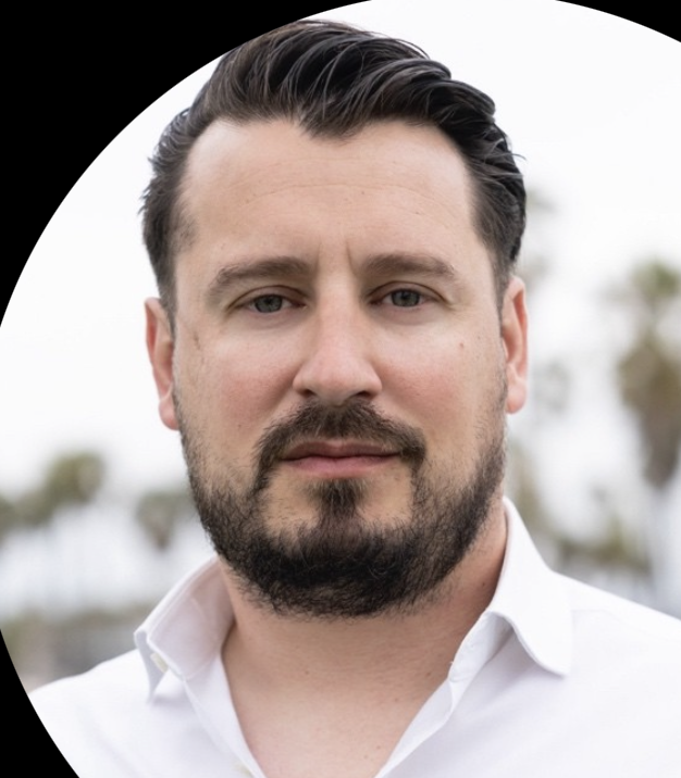 Portrait of a man with dark hair and beard wearing a white collared shirt, with blurred outdoor background.