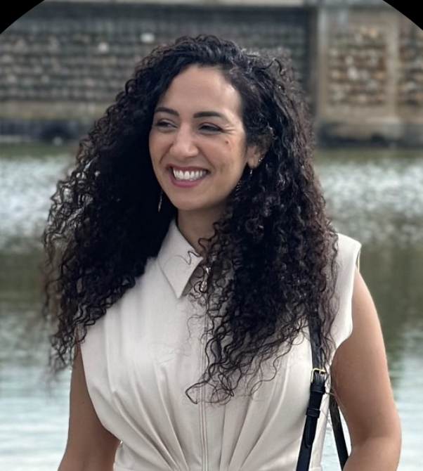 Smiling woman with long curly hair wearing a sleeveless white dress standing near a body of water.