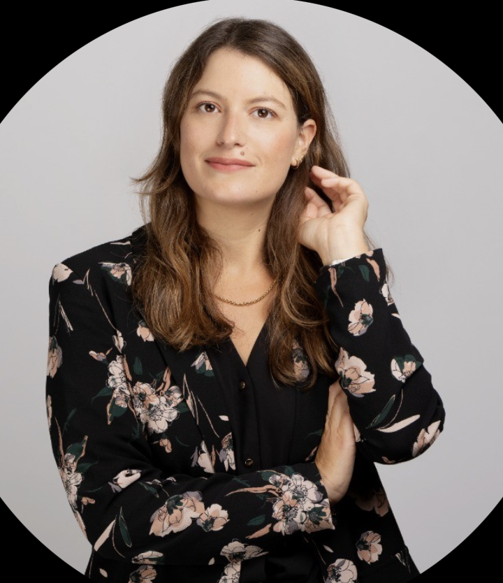 Woman with long brown hair wearing a black floral blazer and a gold necklace, posing against a light gray background.