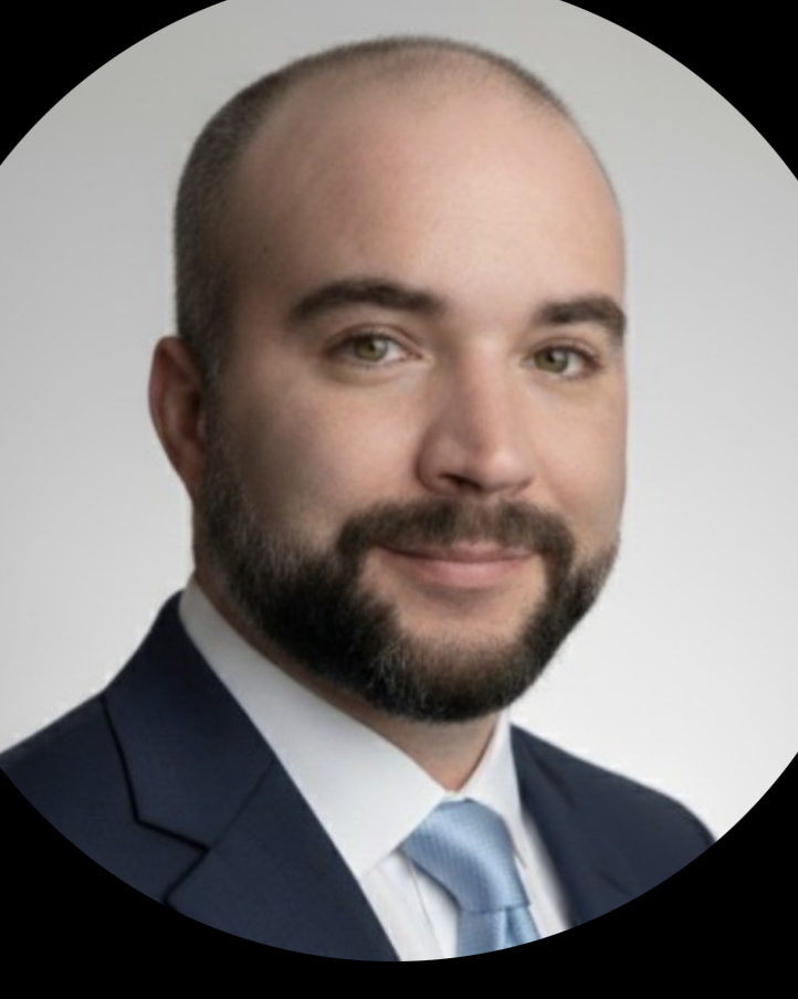 Close-up portrait of a smiling bald man with a beard, wearing a navy suit, white shirt, and light blue tie.