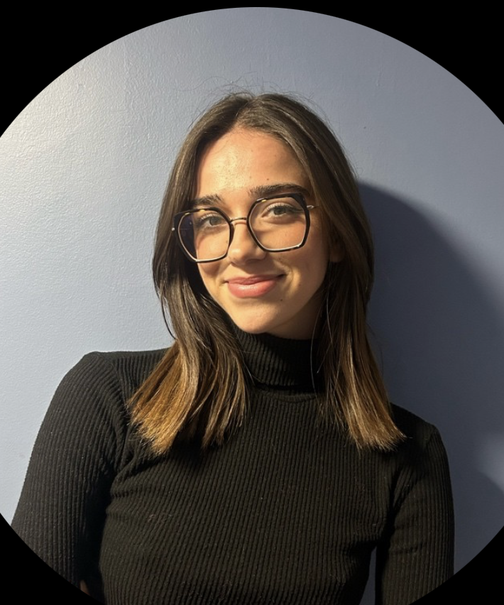 Young woman with shoulder-length brown hair and glasses, wearing a black turtleneck and smiling against a plain light blue background.