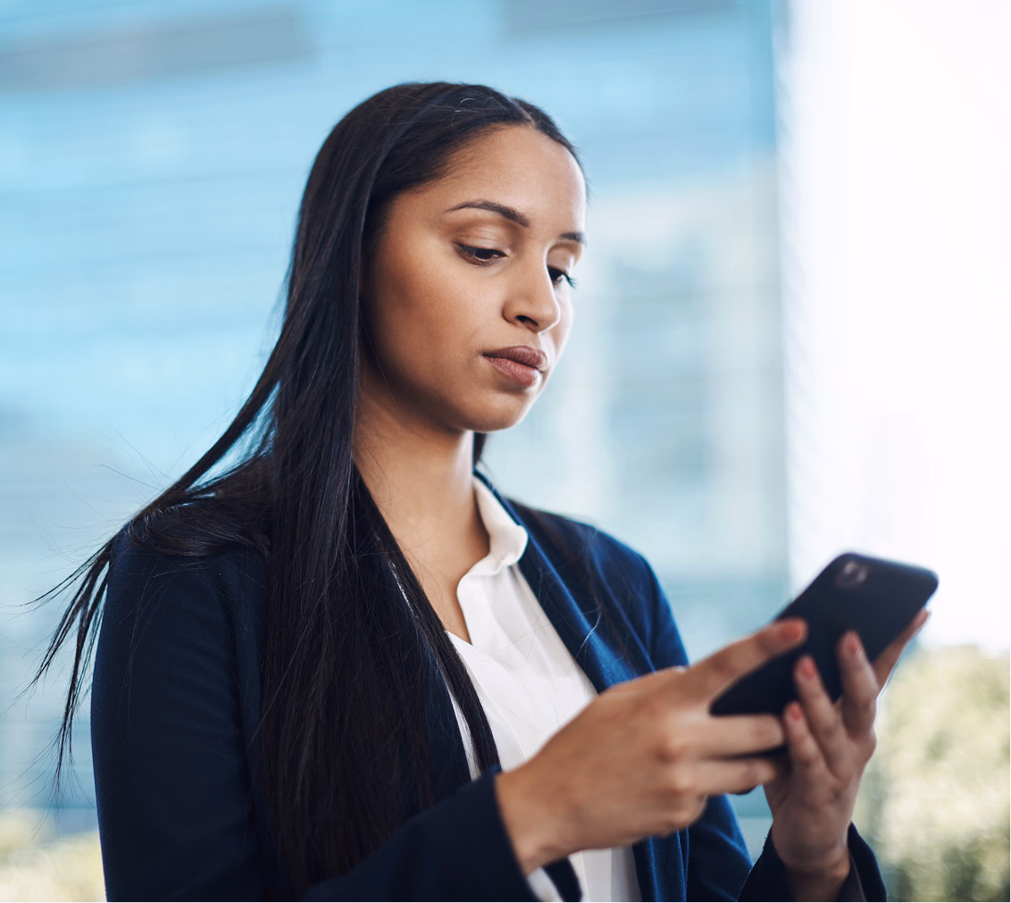 A focused young woman in a blazer holds and looks at a smartphone in an office setting.