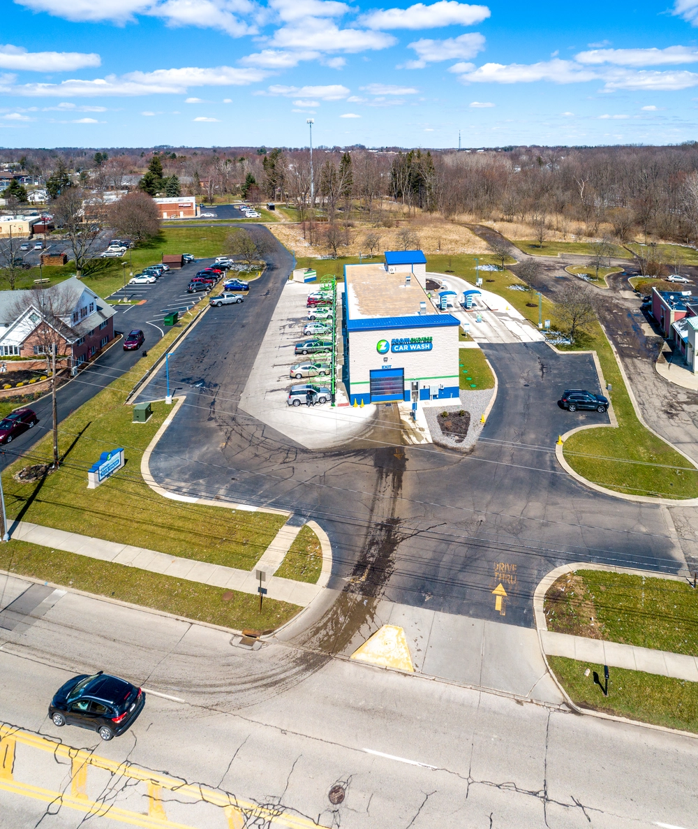 Aerial view of a self-service car wash facility with several cars parked and a clear drive-thru lane under a blue sky with scattered clouds.