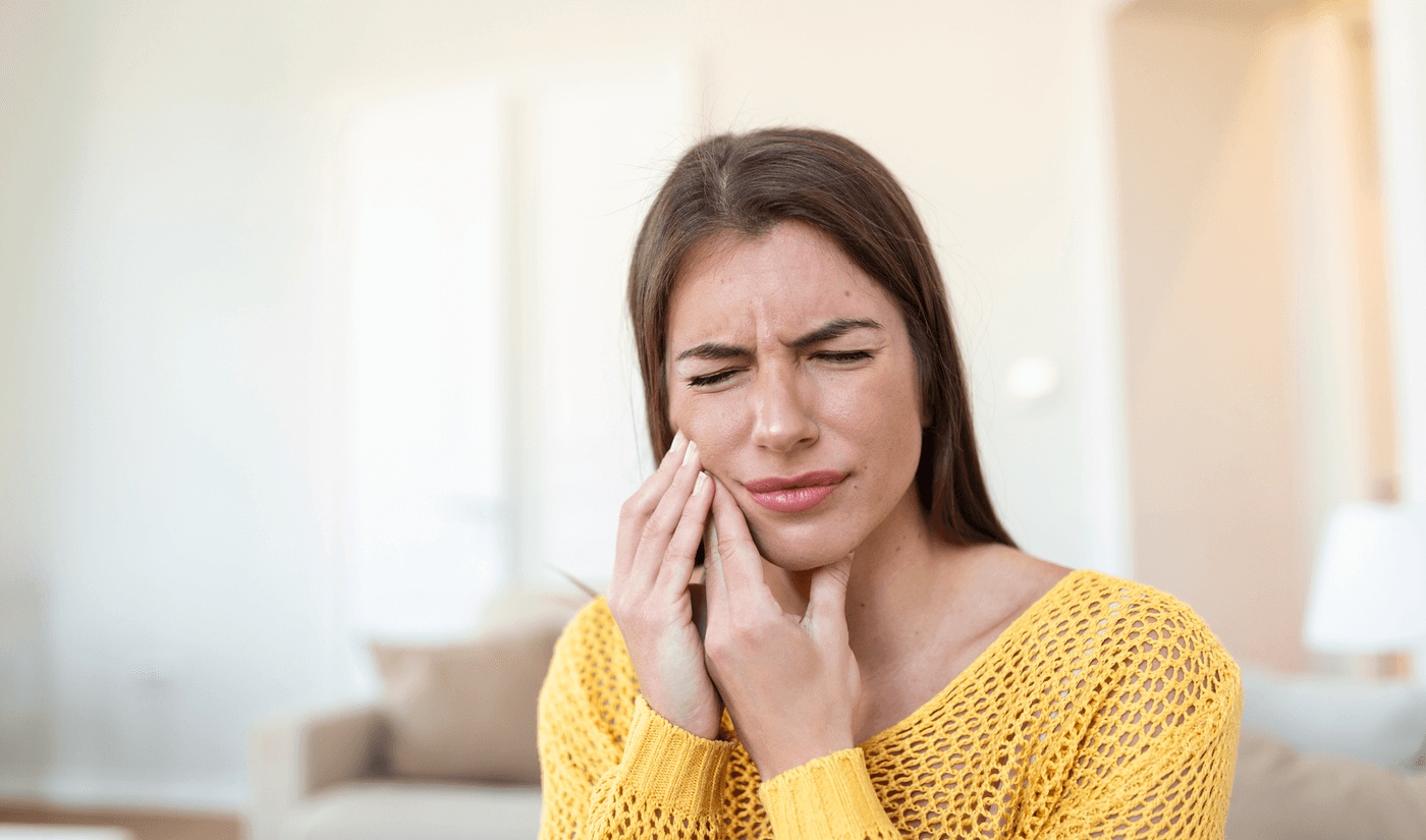 Young woman holding her jaw in pain at home, showing signs of a dental emergency and needing care from the best emergency dentist in Miami Beach.