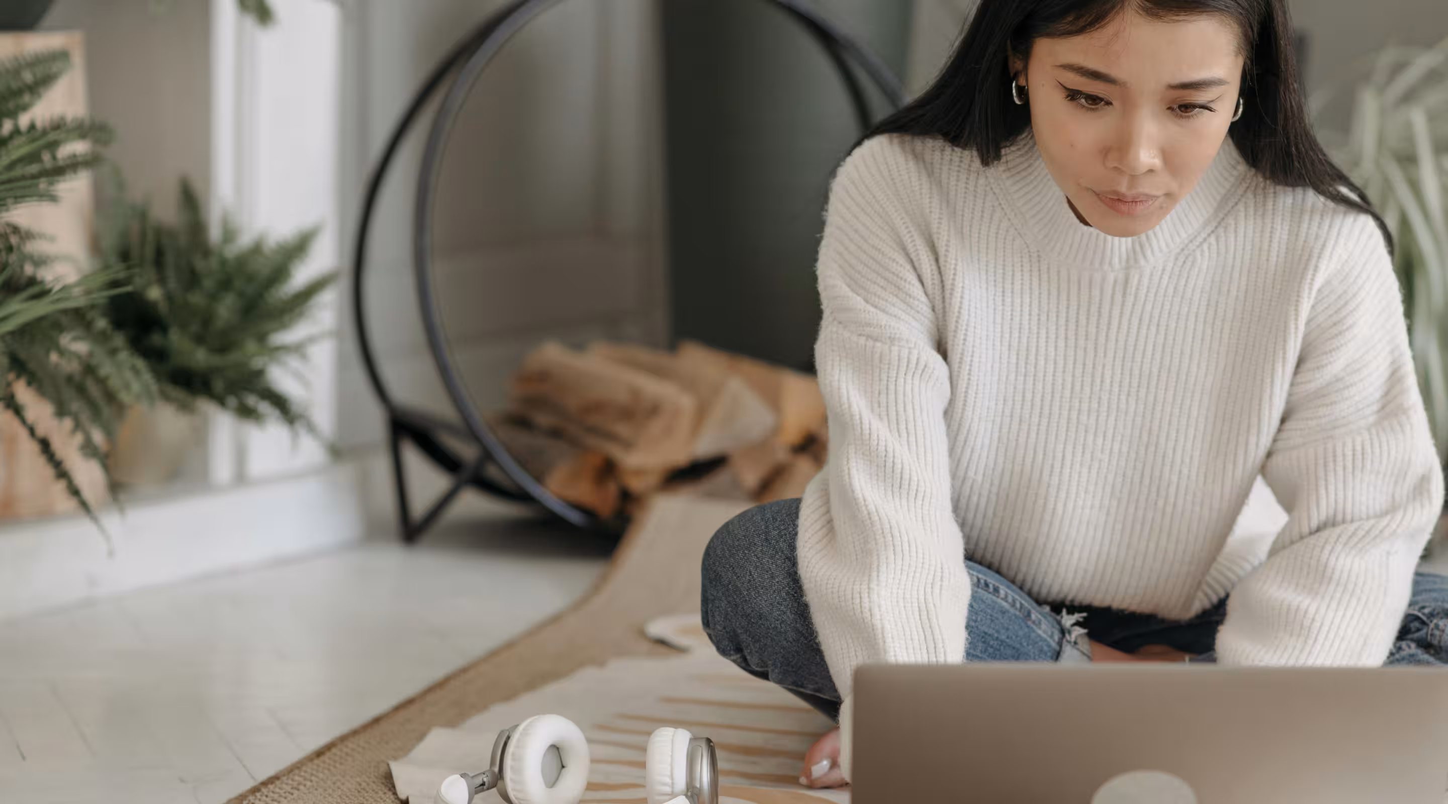 Woman in a white sweater sitting cross-legged on the floor using a laptop to read Emails, with headphones beside her.