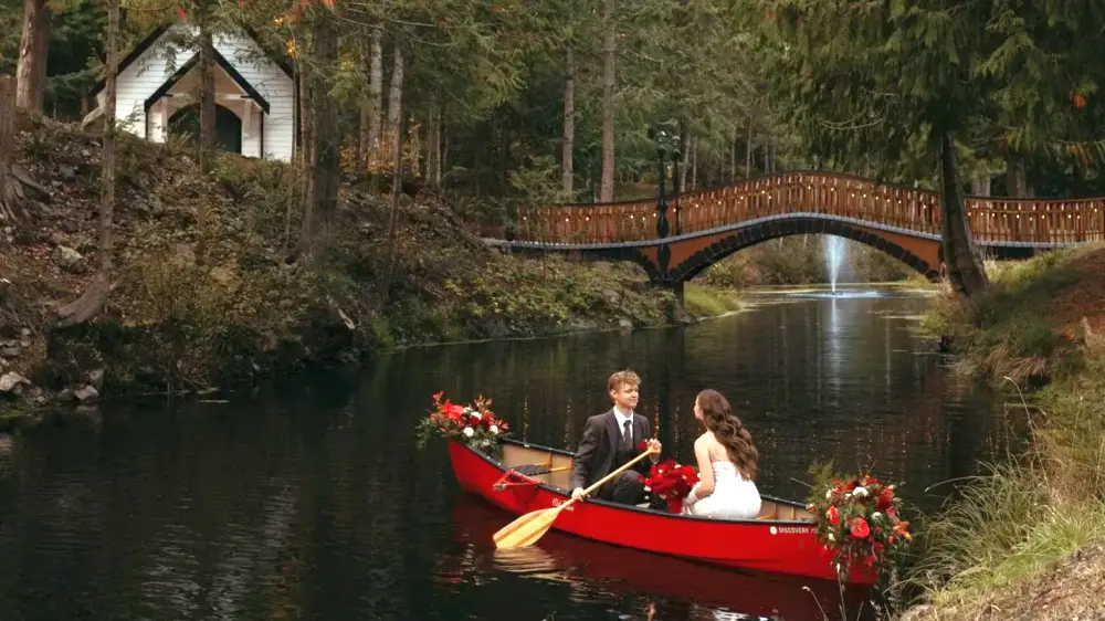 A bride and groom sit in a red kayak decorated with flowers, with a custom built wood bridge and fountain in the background, next to a white country wedding chapel in Anacortes, WA.