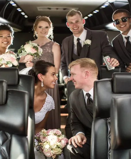 Bride and groom smiling at each other with bridal party holding bouquets inside a limousine.