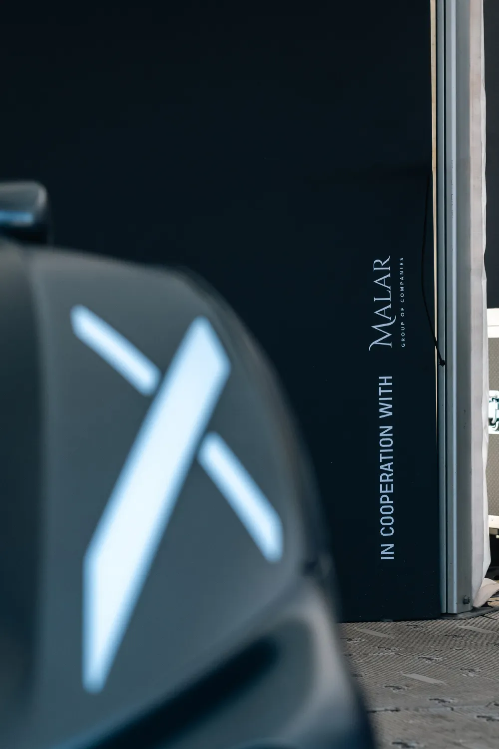 Close-up of the hood of a Bugatti Bolide with a white stylized X logo blurred in the foreground and a black wall in the background with the text 'IN COOPERATION WITH MALAR GROUP OF COMPANIES'.