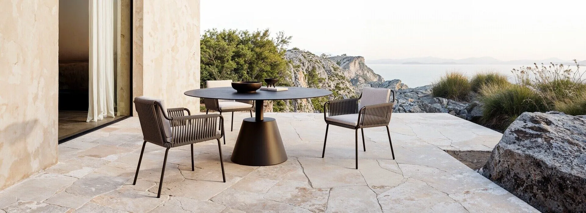Contemporary stone terrace overlooking the sea, featuring outdoor furniture staging with a sleek, round black dining table and modern woven chairs arranged for a coastal dining experience.