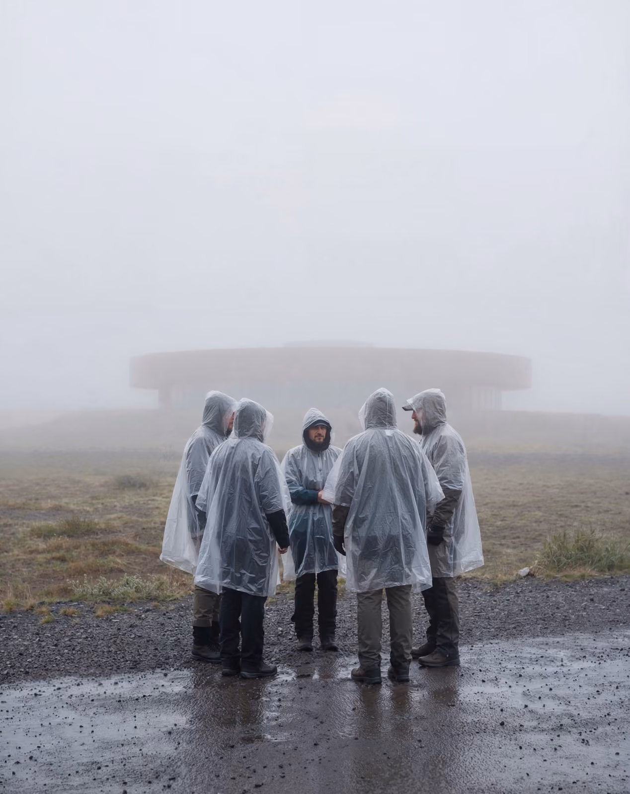 Five people wearing rain ponchos standing on wet ground in foggy weather near a circular building.