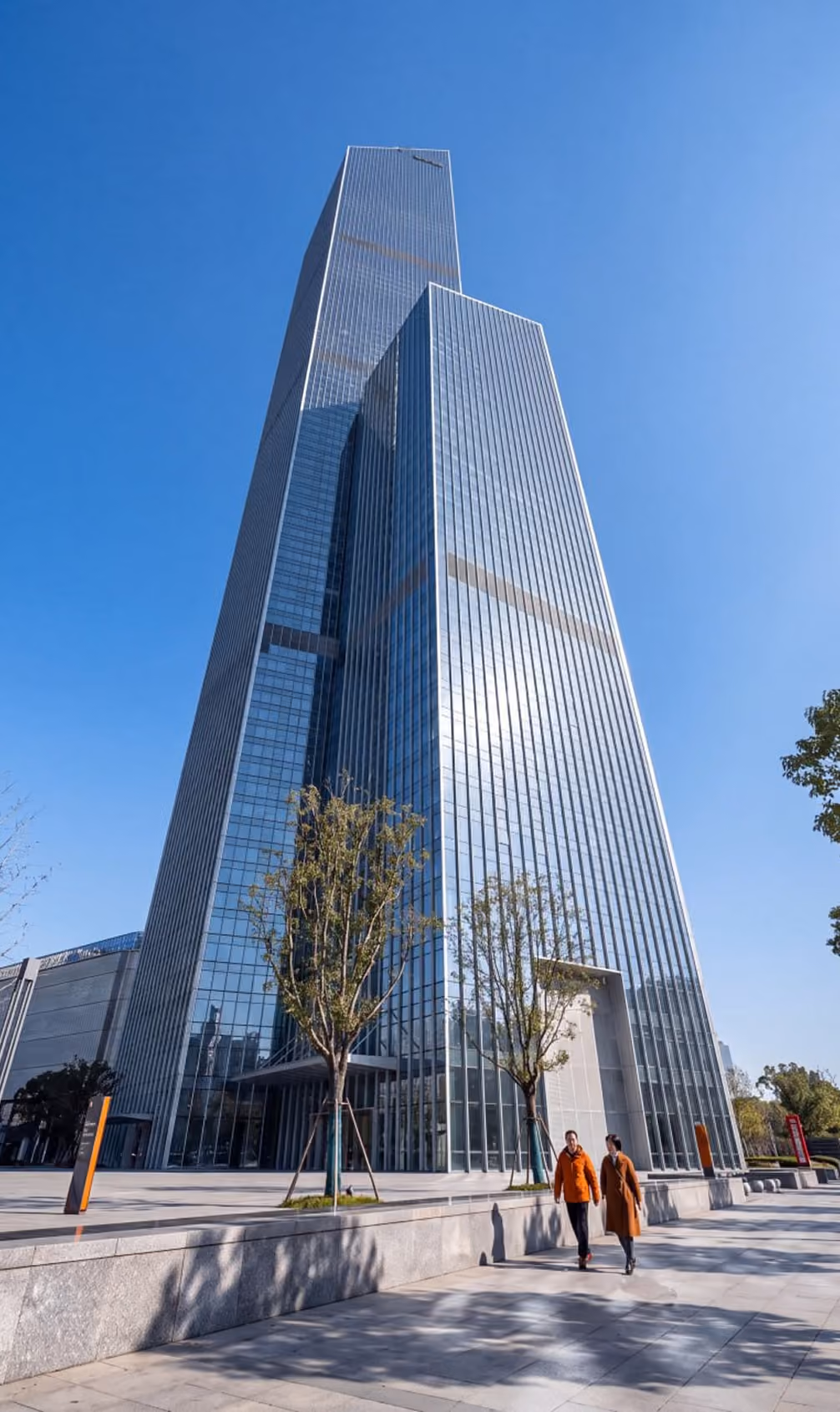 Two people in orange coats walking near tall glass skyscrapers with clear blue sky.