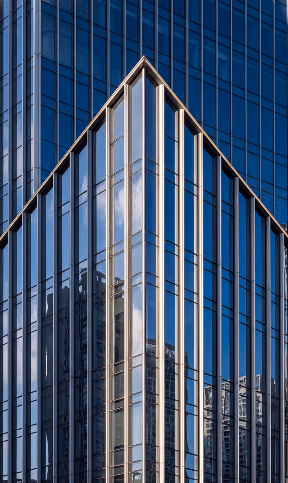 Corner of a modern glass skyscraper reflecting clouds and nearby buildings in blue tones.