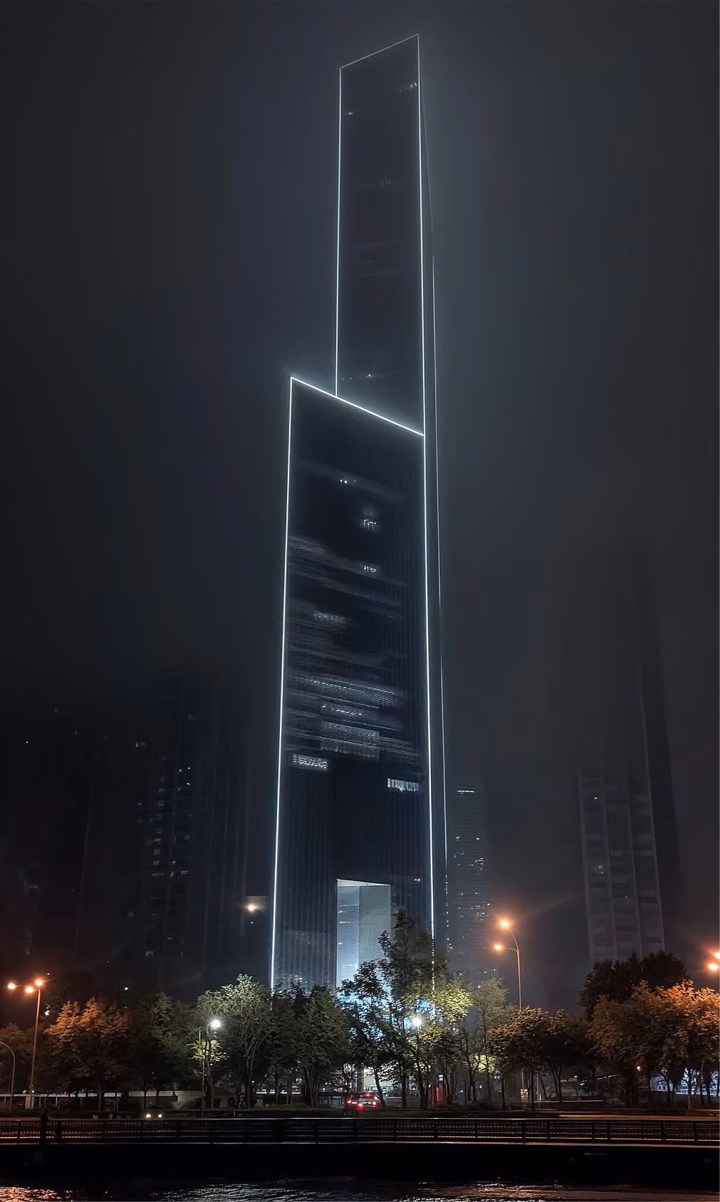 Tall modern skyscraper outlined with bright white lights standing at night with trees and streetlamps below.