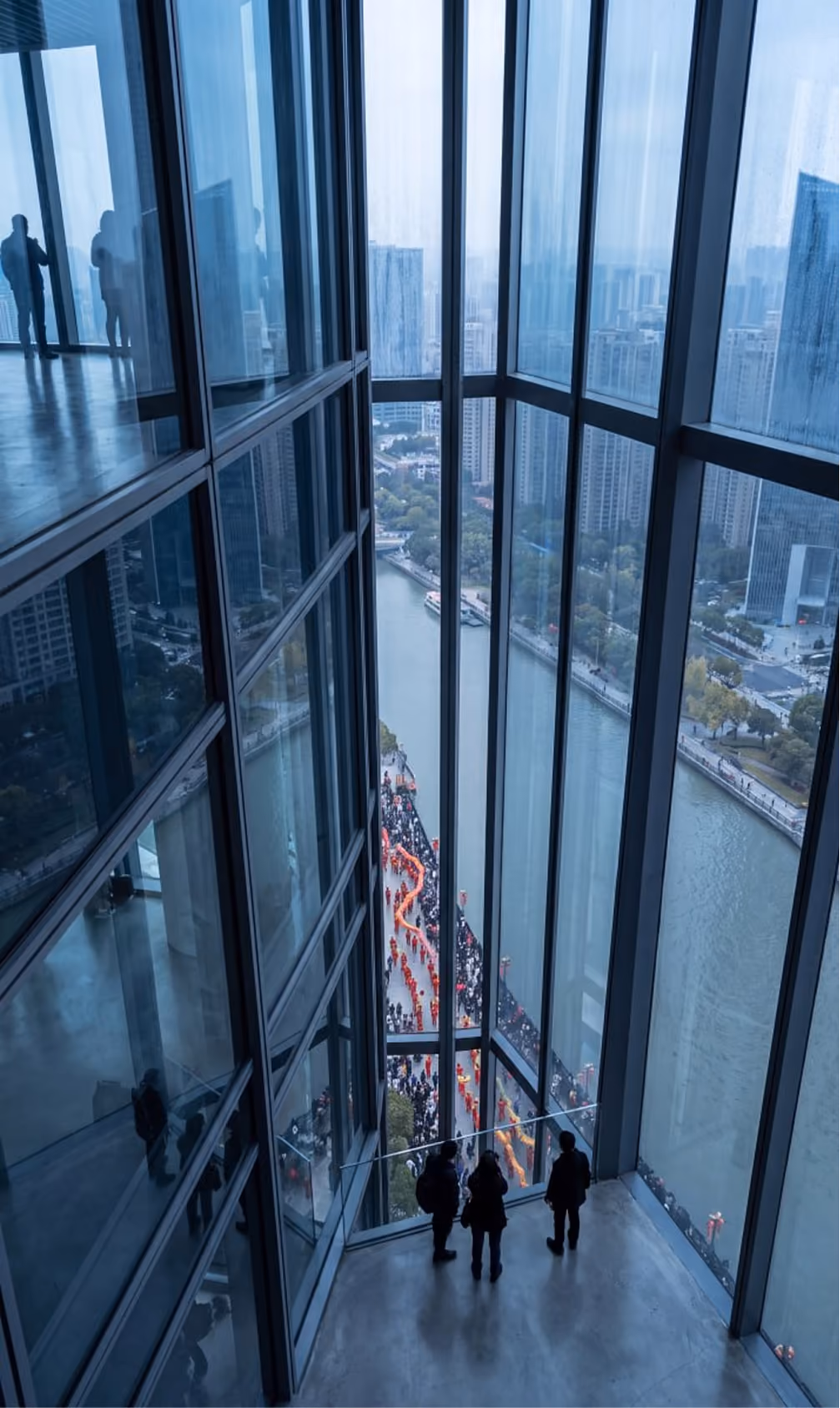 Three people inside a glass building overlooking a river and a crowd with a colorful parade on the street.