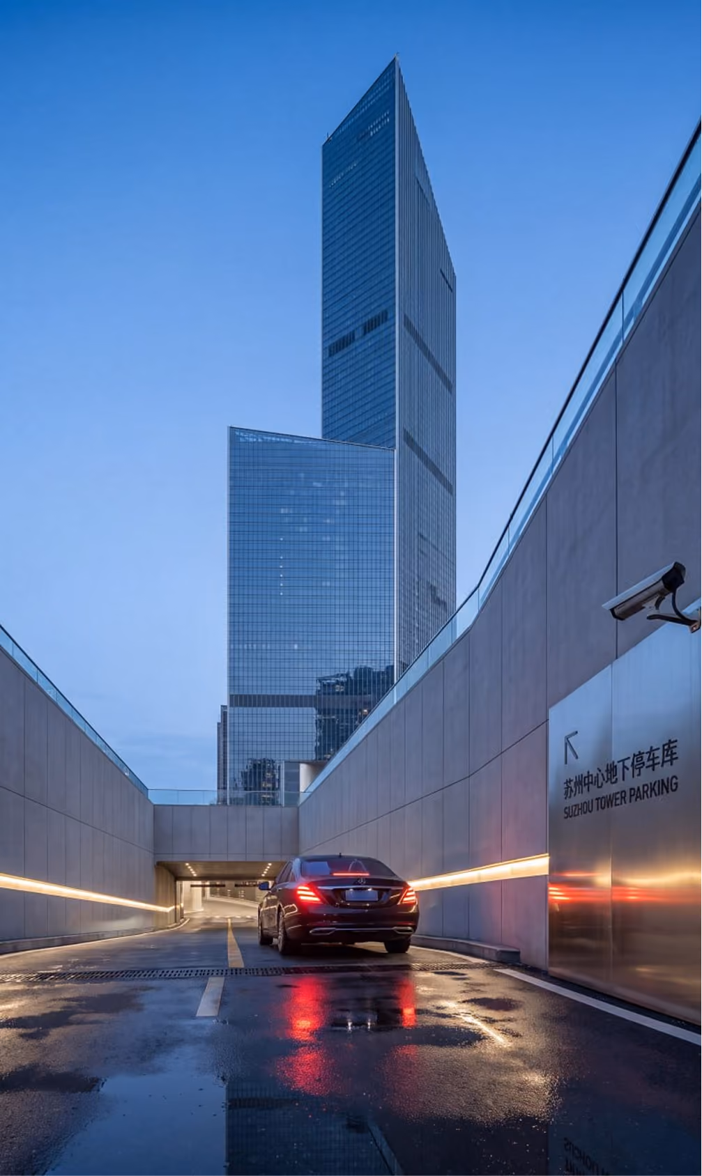 Black sedan driving into the Suzhou Tower parking entrance with tall glass skyscrapers in the background.