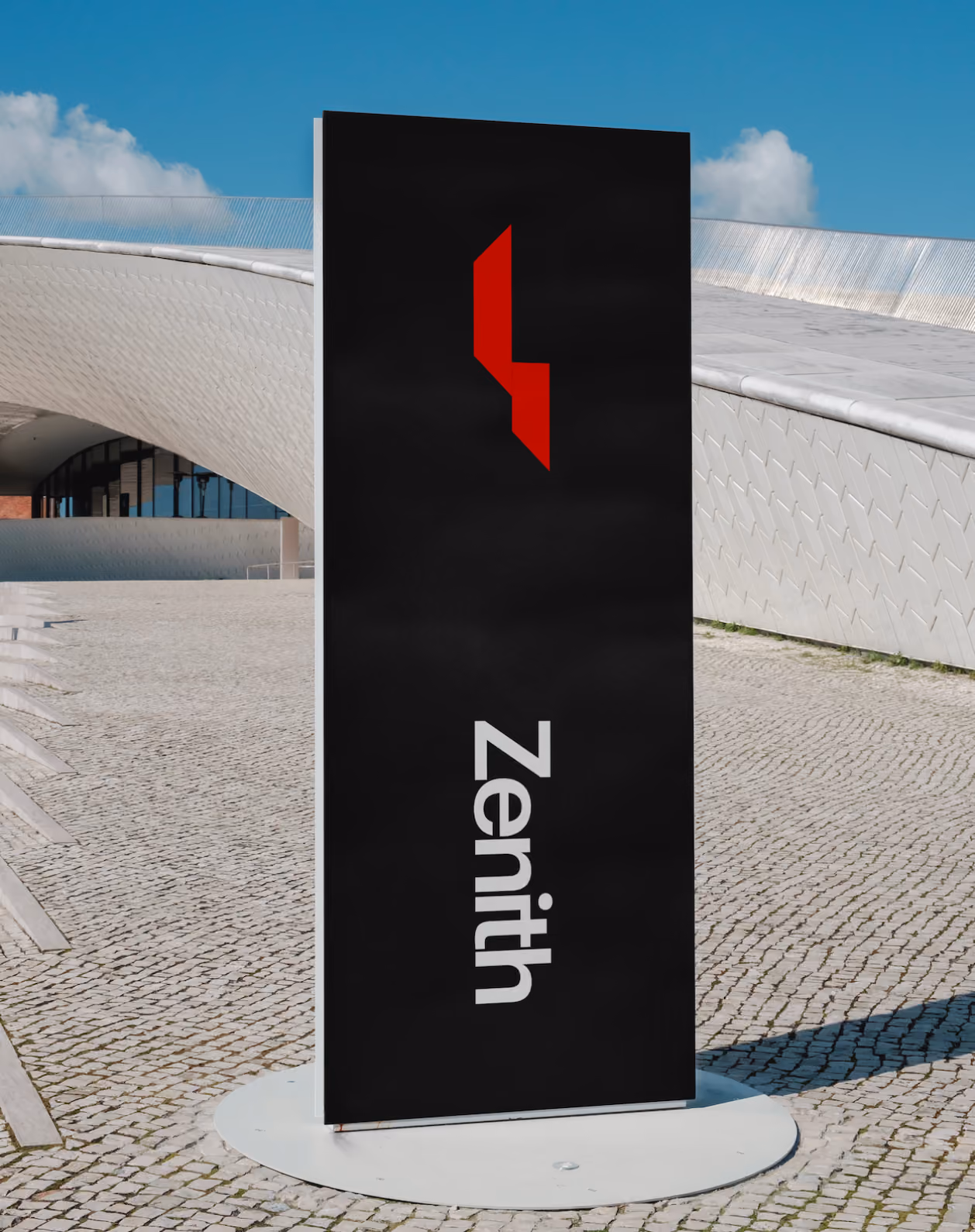 Tall black sign with red geometric logo and white text 'Zenith' on a paved plaza under a blue sky.