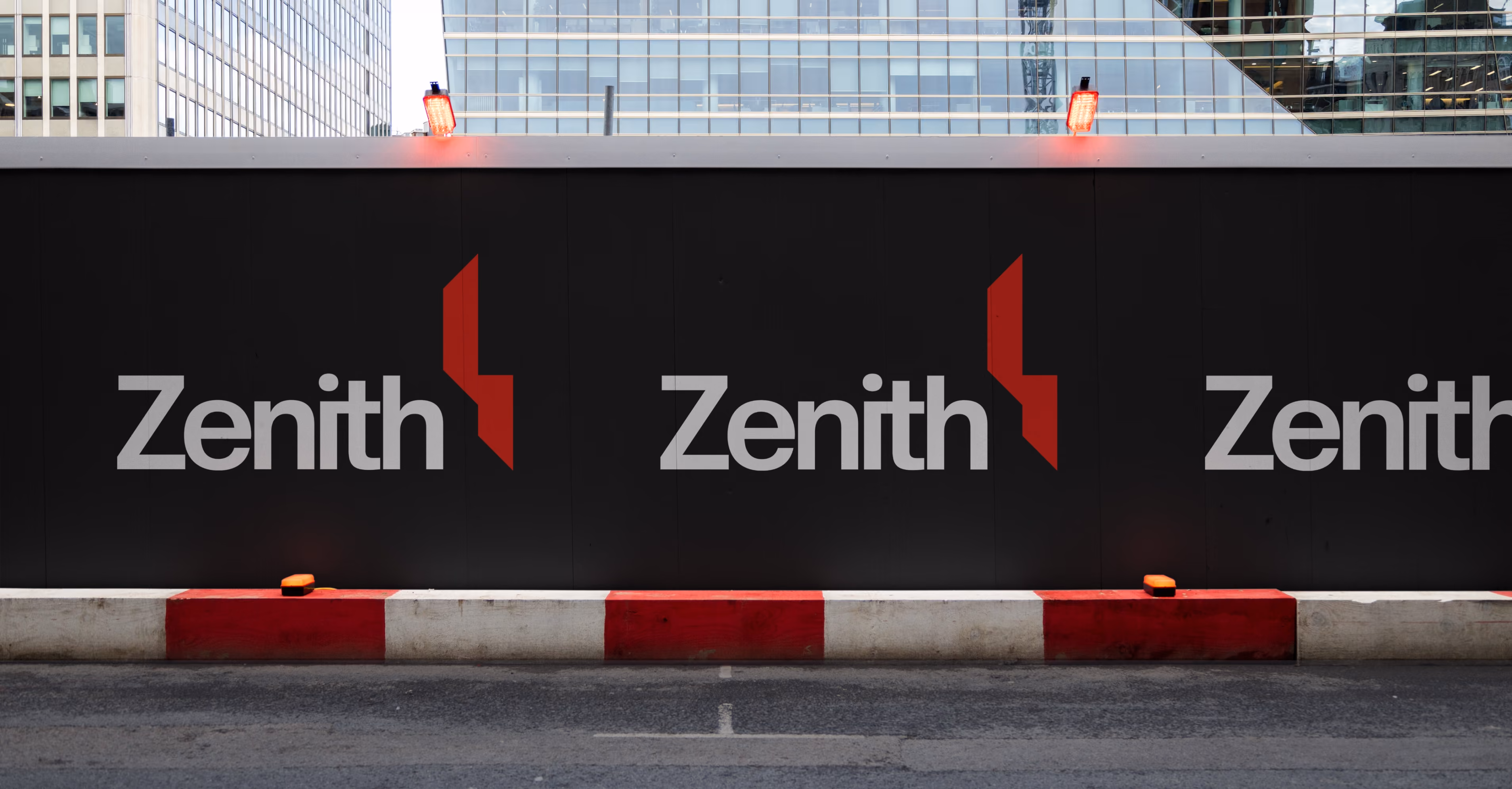 Black construction barrier with repeating white and red Zenith logos and city buildings in the background.