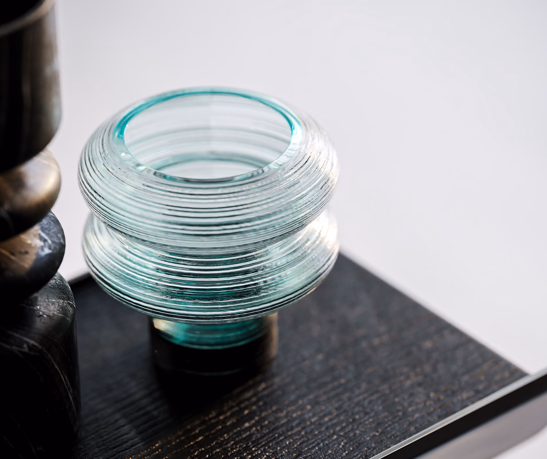 Close-up of a ribbed glass object on a dark wooden surface with parts of a dark sculpture nearby.