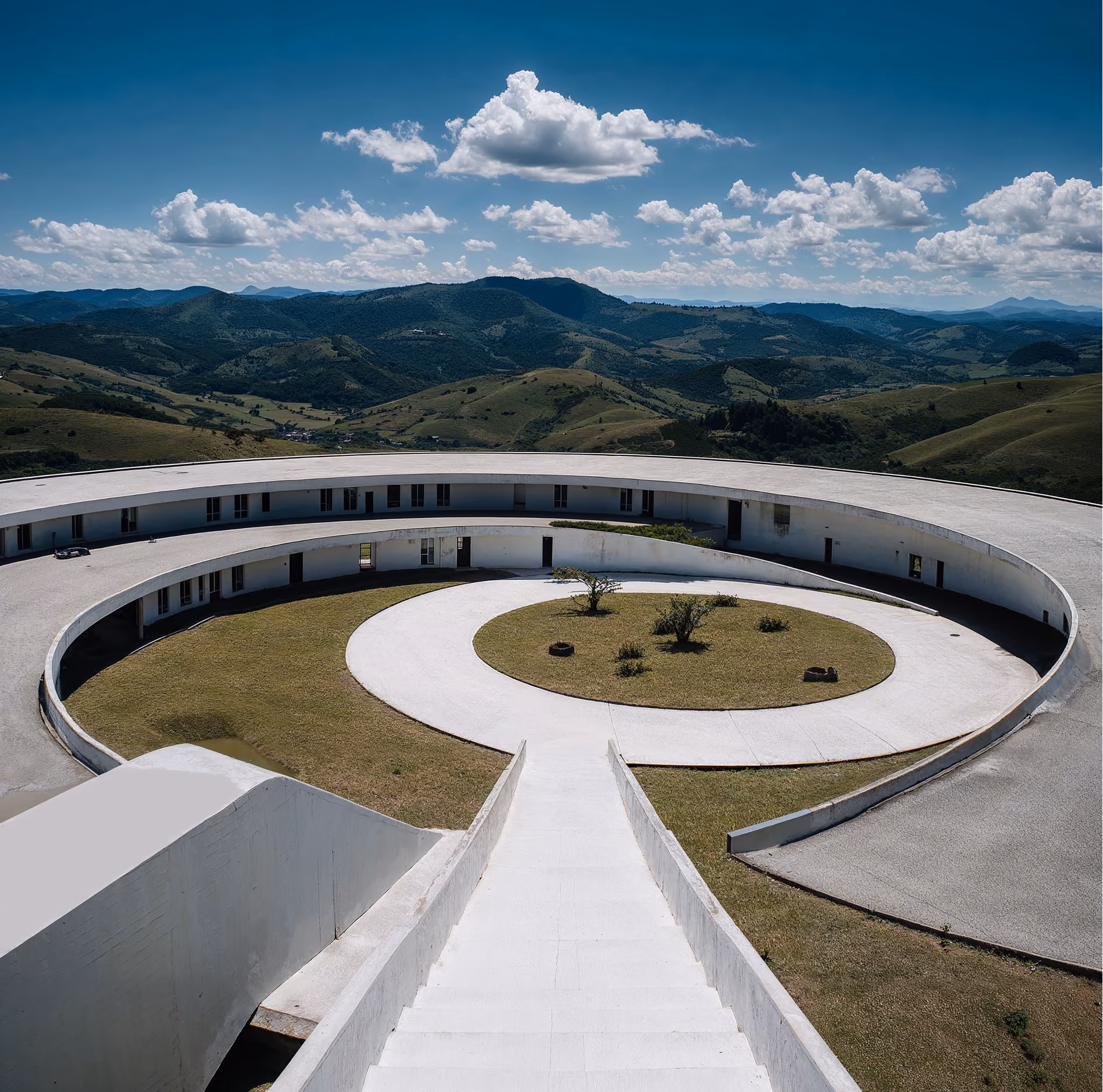 White circular building with inner courtyard and pathway surrounded by green hills under a blue sky.