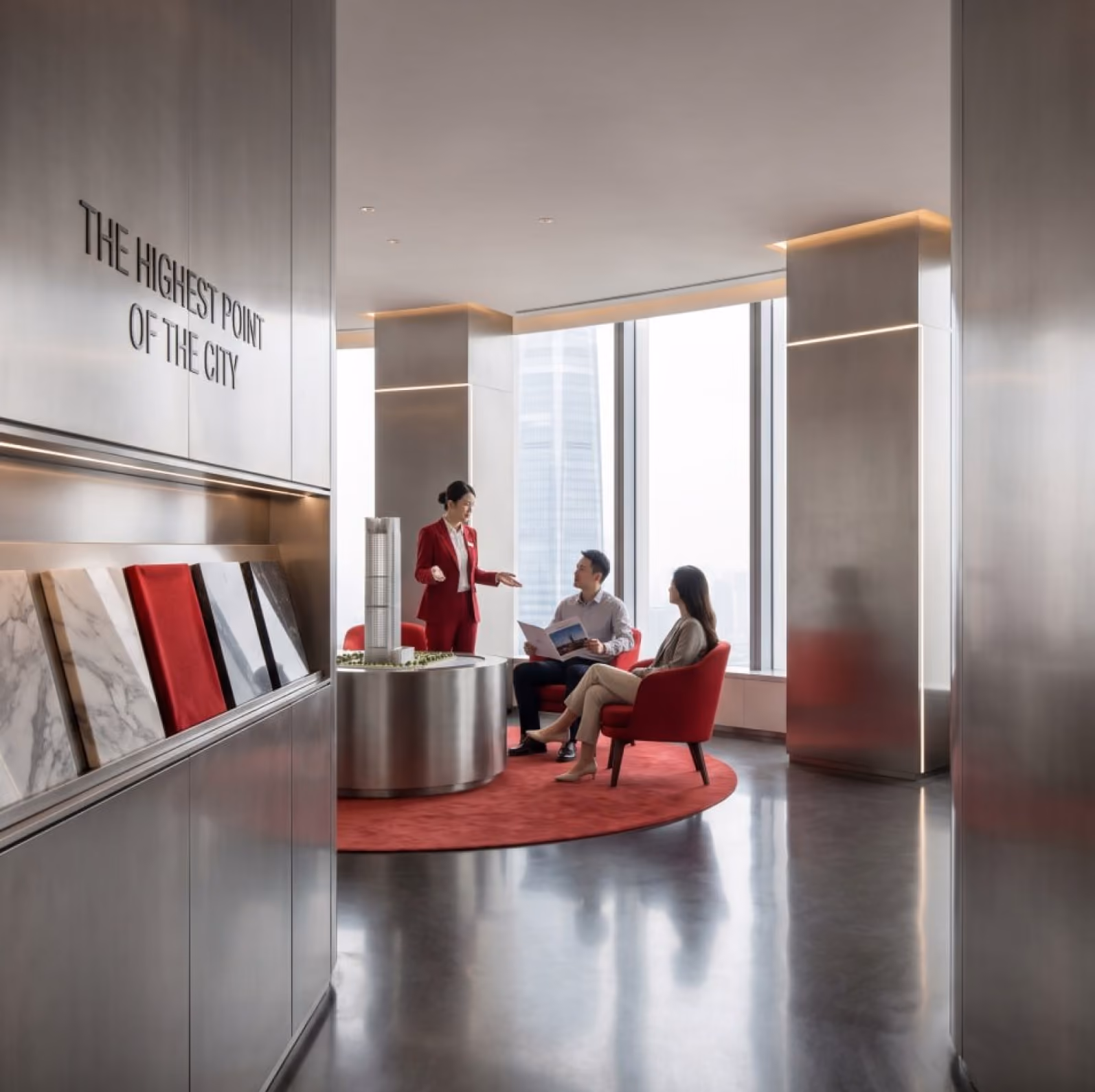 Three people discussing building models in a modern showroom with large windows and red chairs.