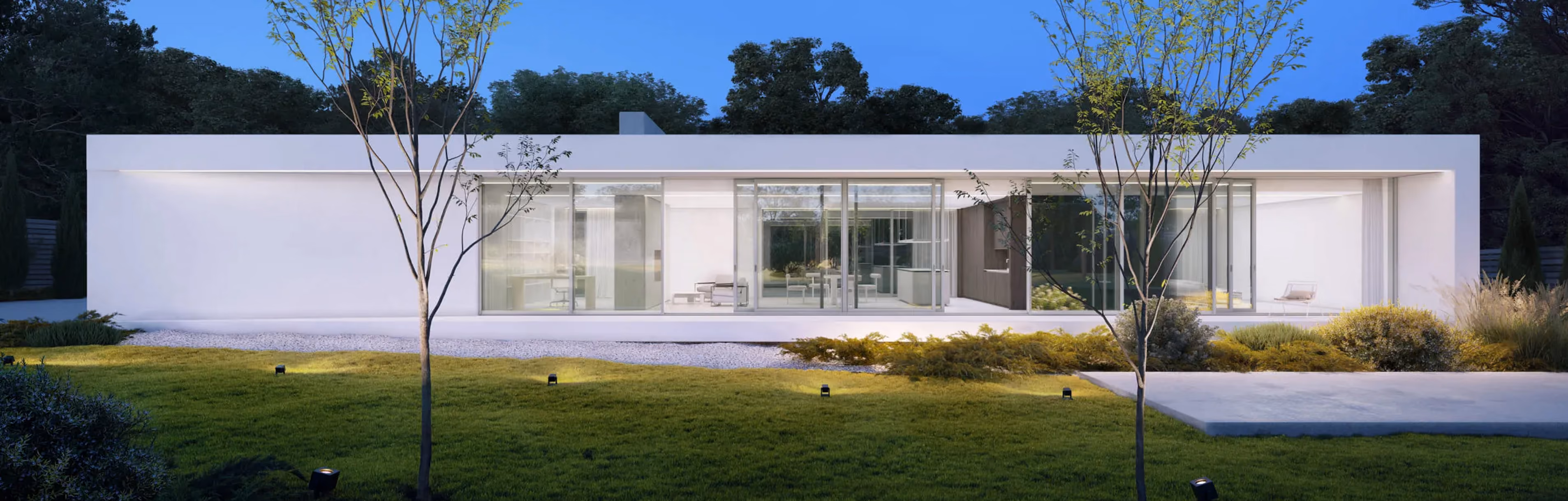 Modern single-story white house with large glass windows and a green lawn with small trees at dusk.