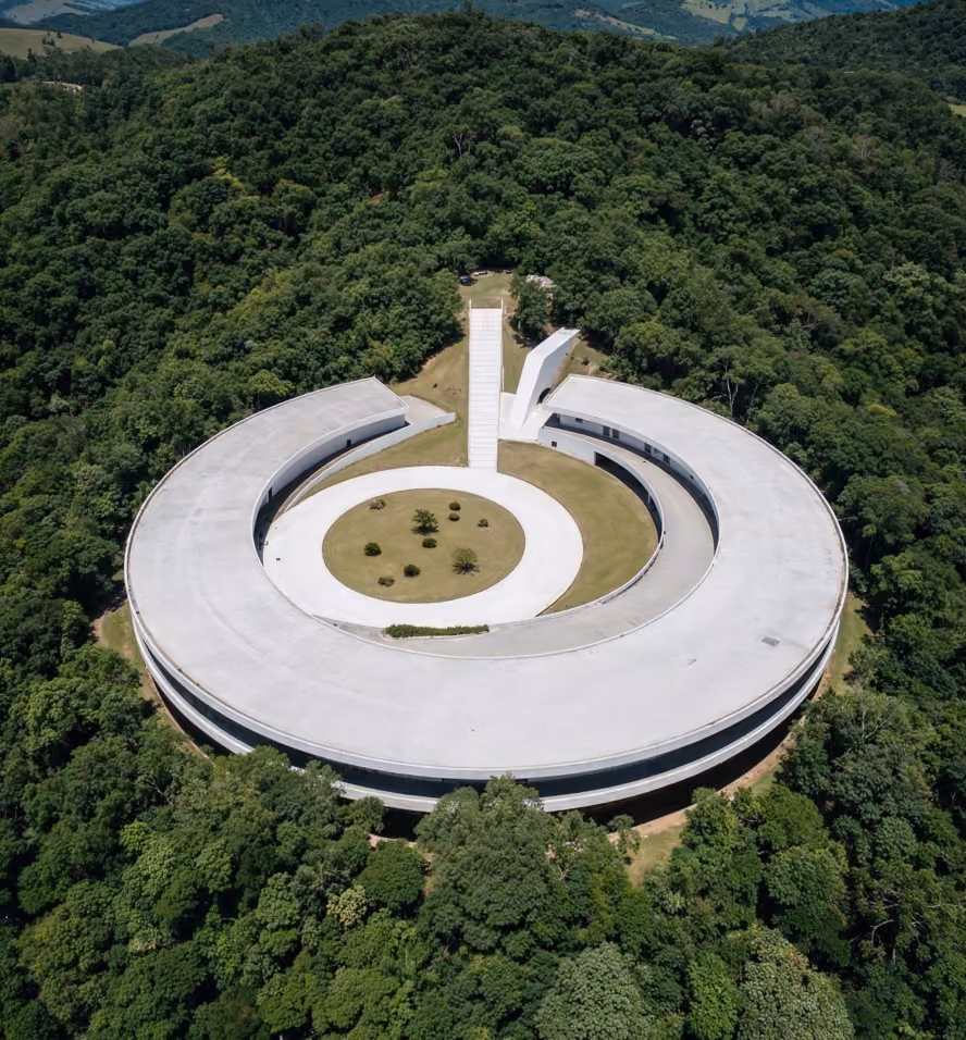 Aerial view of a large, circular, modern building with a central open courtyard containing a lawn and small trees, surrounded by dense forest.