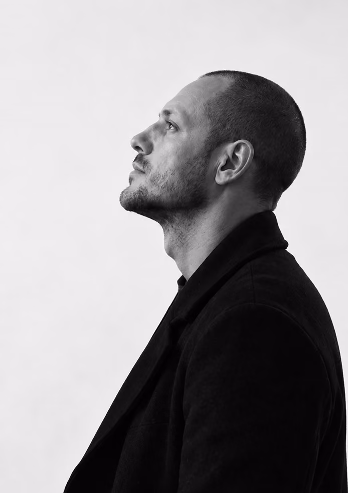 Black and white profile portrait of a man with a short beard and buzzed hair looking upwards, wearing a dark coat against a plain light background.