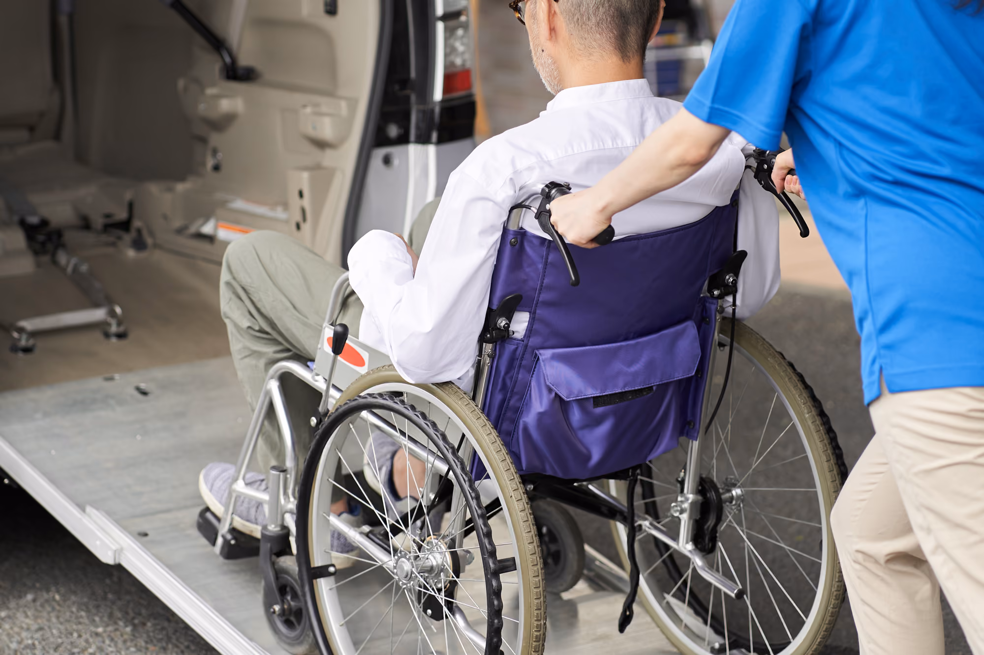 Caregiver in blue scrubs pushing an elderly man in a wheelchair up a ramp into a vehicle.