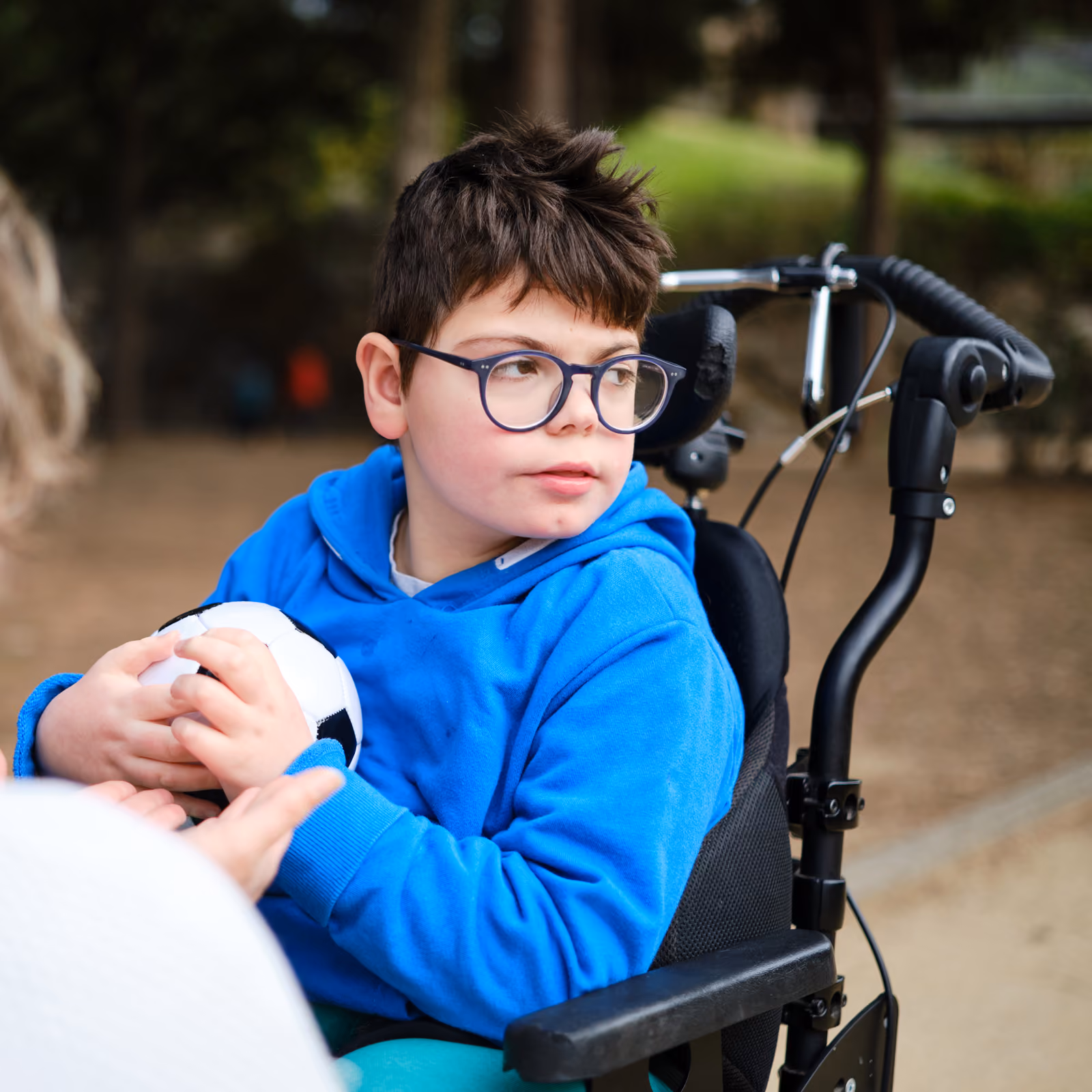 Boy in wheelchair wearing glasses and a blue hoodie holding a small soccer ball outdoors.