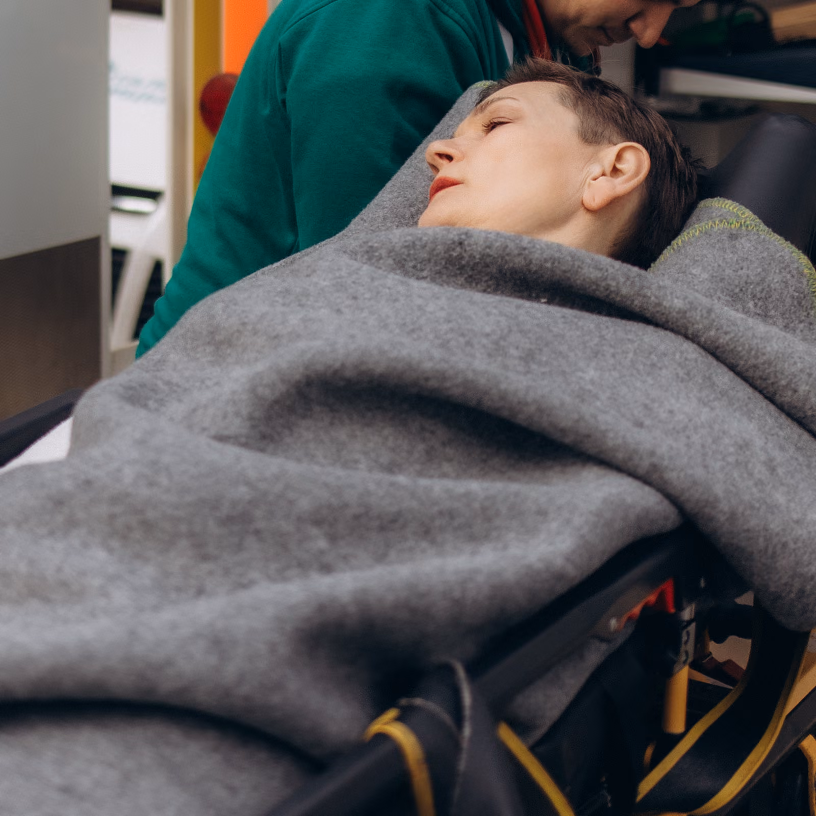 Person resting on a stretcher covered by a gray blanket with another person leaning close.