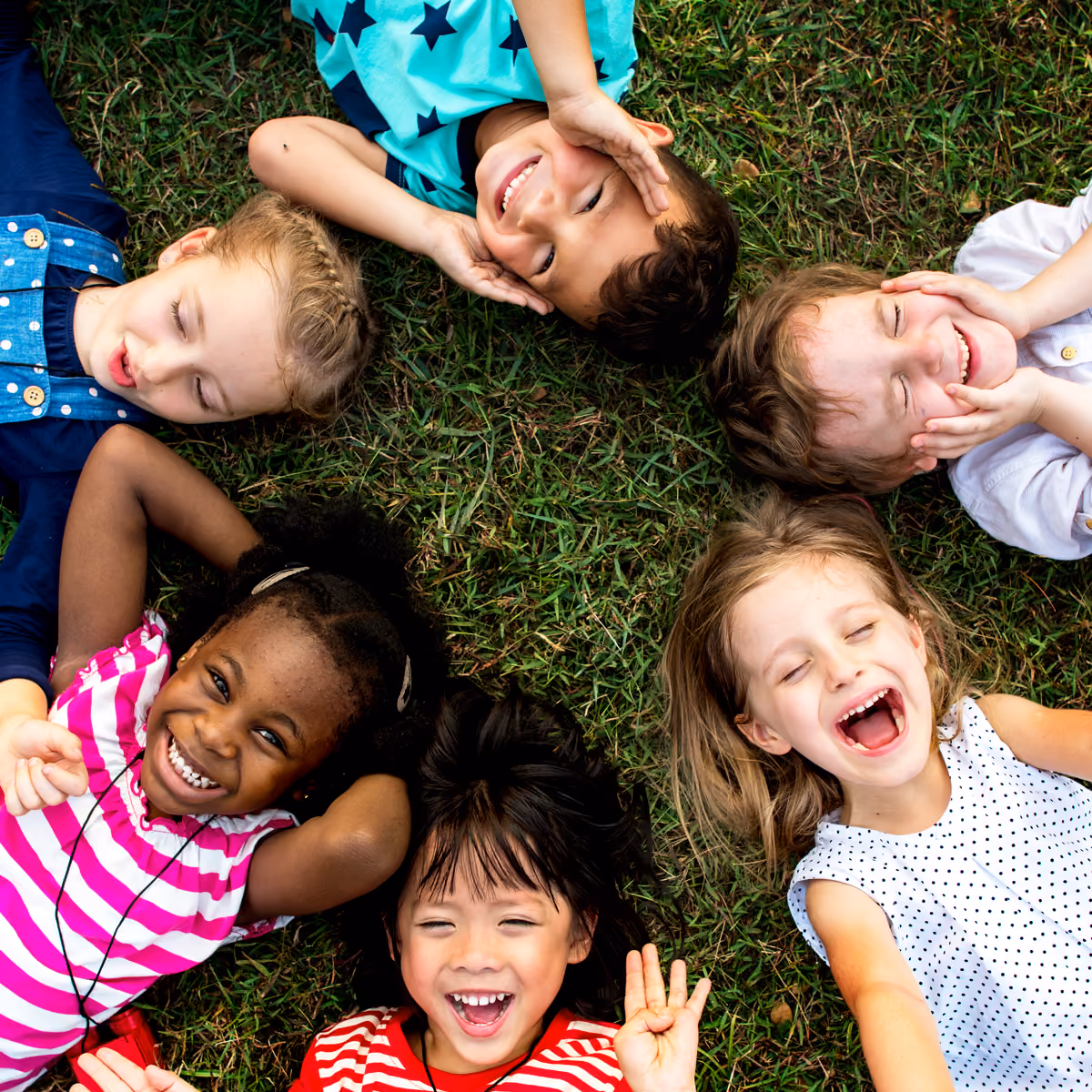 Six diverse children lying in a circle on grass, smiling and laughing with eyes closed or open.