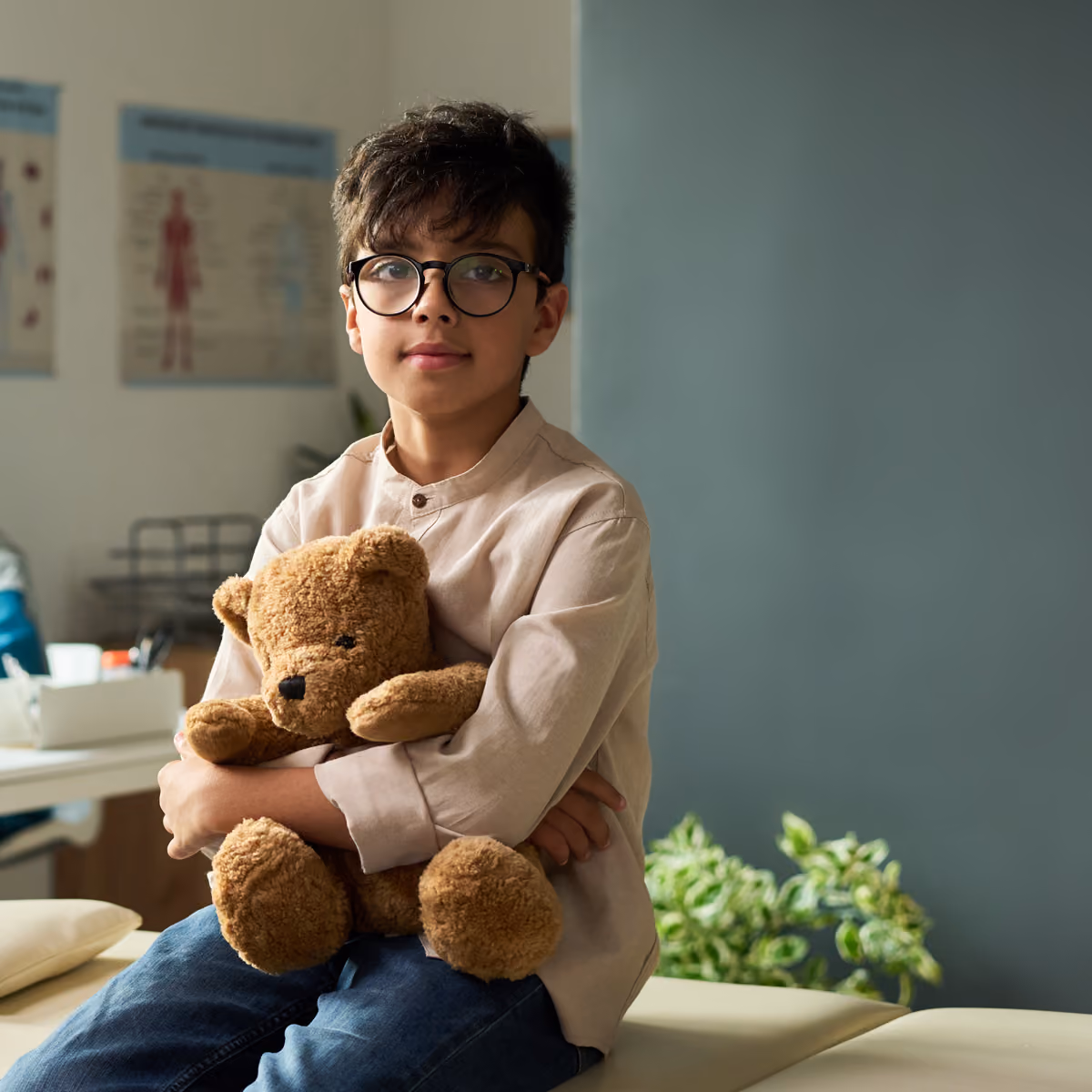 Boy with glasses sits in a waiting room holding a brown teddy bear.