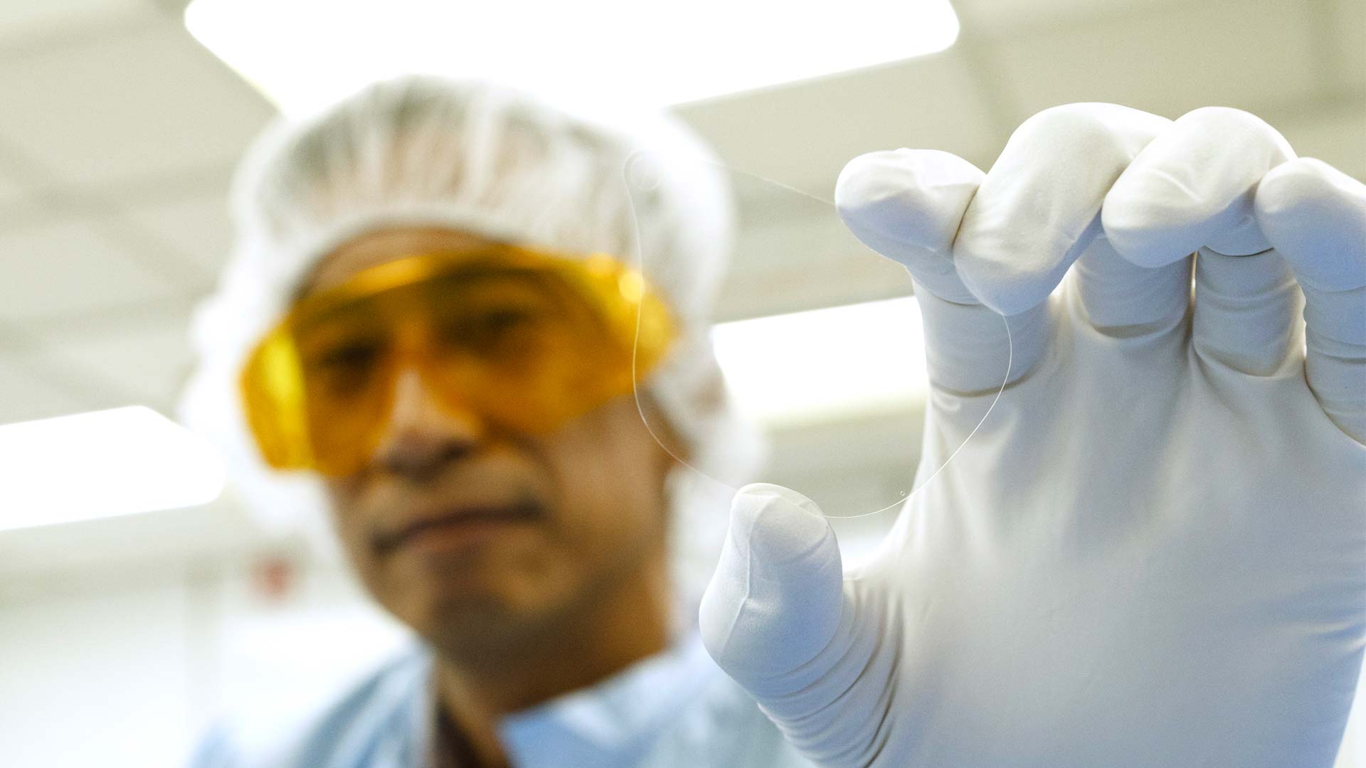 A lab technician holds up a transparent waveguide in his gloved hand.