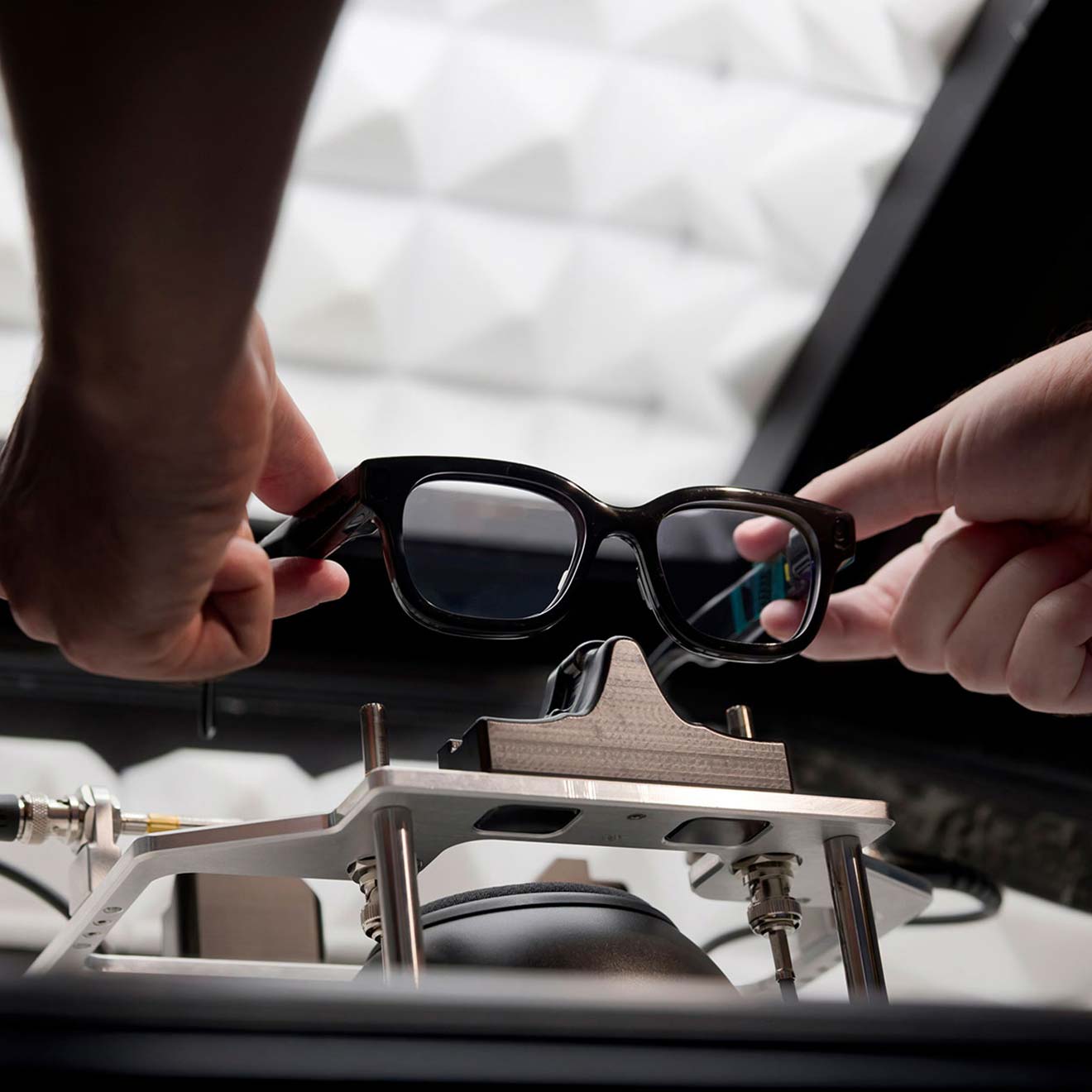 Hands gently setting a pair of AR glasses into a cradle within a brightly lit testing chamber.