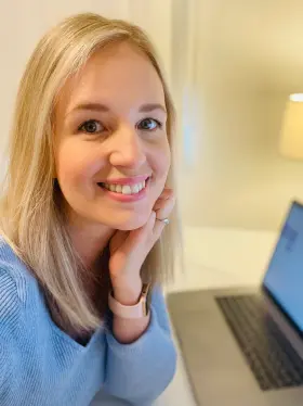 Nainen istuu työpöydän ääressä tietokone avonaisena edessään ja hymyilee kameralle.