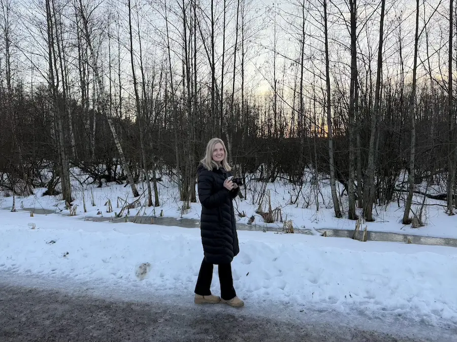 Nainen seisoo talvisen maiseman edessä kamera kädessään, katsoo kameraan ja hymyilee.