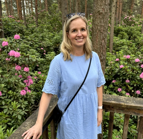 Nainen seisoo sinisessä kesämekossa rhododendrojen ympäröimänä hymyillen ja katsoen kameraan.