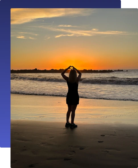 Nainen seisoo hiekkarannalla selin kameraan auringonlaskun aikaan ja muodostaa käsillään sydämenmerkin.