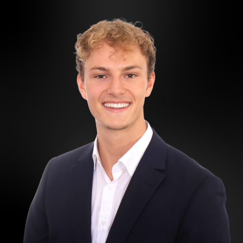 Junger Mann mit lockigem blonden Haar, weißem Hemd und dunklem Sakko vor schwarzem Hintergrund.