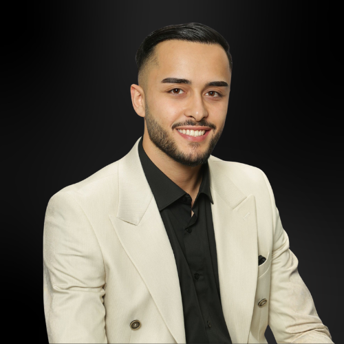 Ein junger Mann mit kurzem schwarzen Haar und Bart trägt ein cremefarbenes Sakko und ein schwarzes Hemd, lächelt vor schwarzem Hintergrund.